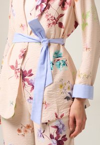 Haut enroulé à imprimé floral dans un tissu texturé pêche, avec une ceinture nouée bleu clair et des poignets, arborant diverses fleurs colorées dans des teintes de rose et de bleu.