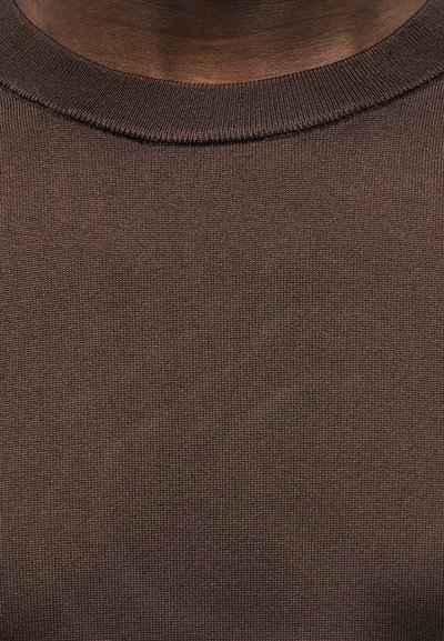 Κοντινό πλάνο στον λαιμό και το πάνω μέρος του στήθους ενός ατόμου που φοράει καφέ πλεκτό πουλόβερ με στρογγυλή λαιμόκοψη.