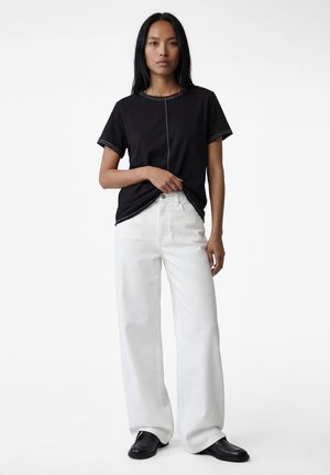 Femme aux longs cheveux noirs portant un T-shirt noir à manches courtes, un pantalon blanc à jambes larges et des chaussures noires, debout devant un fond blanc.