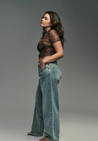 Brūnas leoparda raksta cropped tops ar īsām piedurknēm, apvienots ar brīvām, gaišām augsta jostasvieta džinsām, uz modeļa pret pelēku fonu.