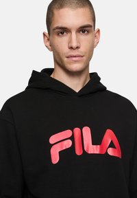 Joven con el cabello corto, vistiendo una sudadera negra con capucha y un gran logo rojo de FILA en el pecho, mirando al frente.