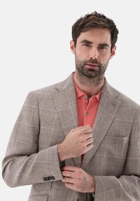 Hellbrauner kariertes Blazer mit subtiler Textur und Reverskragen, getragen über einem korallenfarbenen Poloshirt mit zwei Knöpfen, die Hände des Models passen die Jacke an.