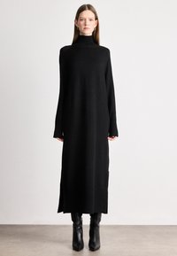 Μαύρο πλεκτό φόρεμα με ψηλό λαιμό και μακριά μανίκια, καθώς και πλευρικές σχισμές. Συνδυασμένο με μαύρες μπότες μέχρι το γόνατο με τακούνι.