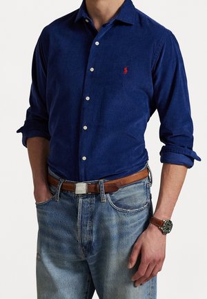 Man wearing a blue button-up shirt with rolled sleeves, light denim jeans, brown leather belt, and brown wristwatch with green dial.