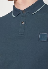 Gros plan sur une personne portant un polo bleu avec deux boutons et un petit patch logo "BOSS" sur la poitrine.