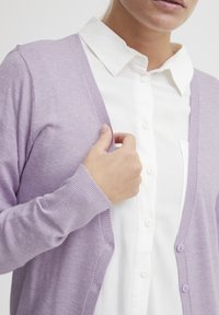 Cardigan lilas clair en tissu tricot doux, avec une fermeture à boutons sur le devant et des poignets côtelés, porté par-dessus une chemise blanche à col.