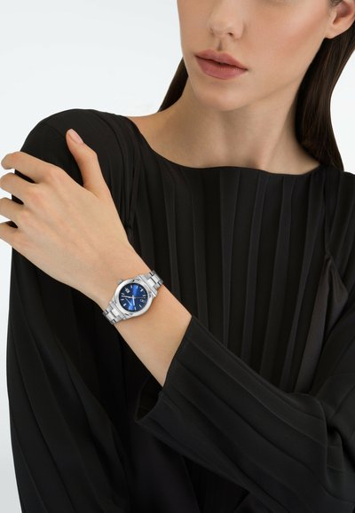 Femme portant un haut plissé noir avec une montre en métal argenté dotée d'un cadran bleu à son poignet gauche, main reposant sur son épaule droite.