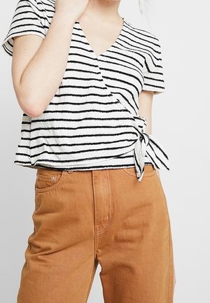 Vrouw draagt een wit-zwart gestreepte wikkel top geknoopt aan de taille met hoog getailleerde bruine broek, hand bij de nek.