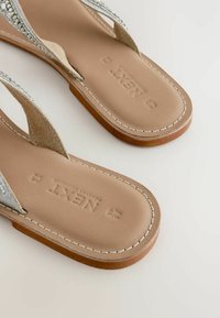 Flip-Flops mit einer braunen Sohle, leichten Wildlederriemen und dekorativen silbernen Verzierungen im oberen Bereich. Genähte Details an den Kanten.