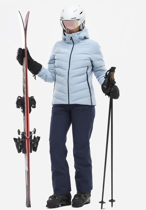 Veste de ski isolante bleu clair avec des accents noirs, pantalon de ski marine, gants noirs, casque blanc et skis avec des détails rouges. Bottes de ski noires.