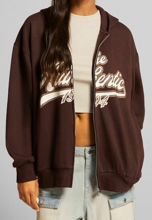 Jeune personne portant un sweat à capuche marron zippé avec le texte "Authentic 1994" sur un crop top blanc et un pantalon cargo bleu clair.