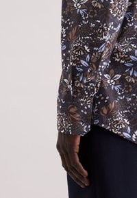 Camicia a fantasia floreale di colore blu scuro, con fiori bianchi, marroni e azzurri; maniche lunghe con polsini a bottoni. Tessuto morbido.