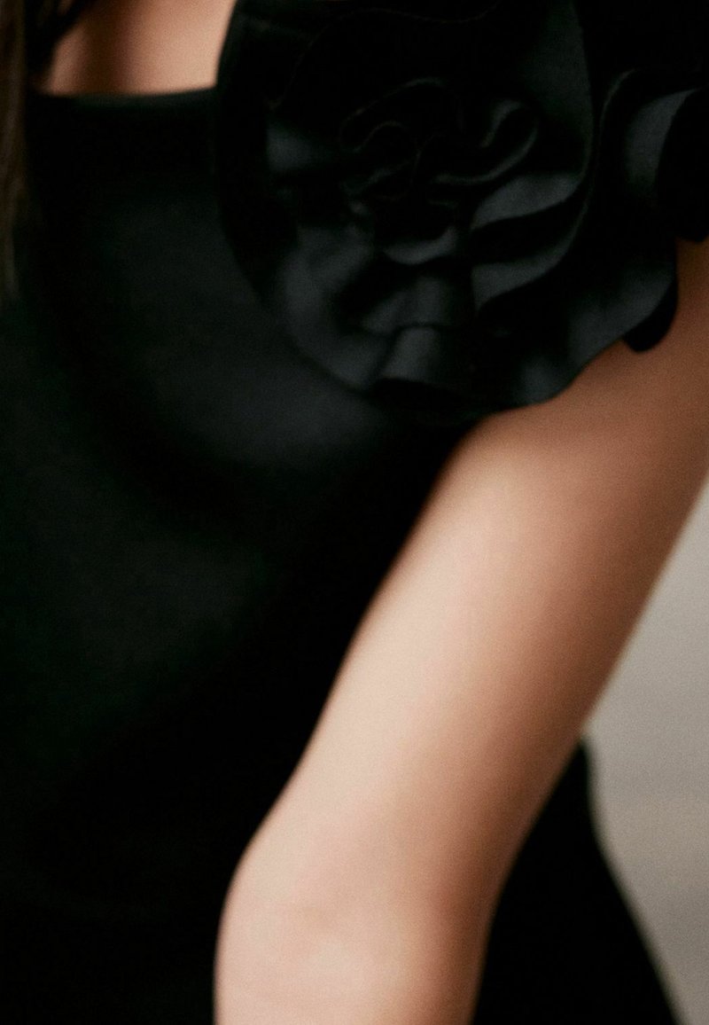 Robe noire avec un détail en tissu à volants sur une épaule. Vue en gros plan montrant la texture lisse et le bras sur un fond neutre.