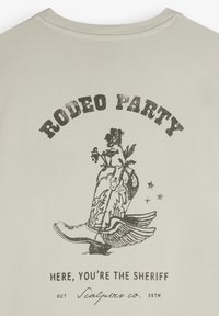 Helles beiges T-Shirt mit einem schwarzen Cowboy-Stiefel-Graphik und floralen Akzenten. Der Text lautet "RODEO PARTY" und "HIER BIST DU DER SHERIFF."