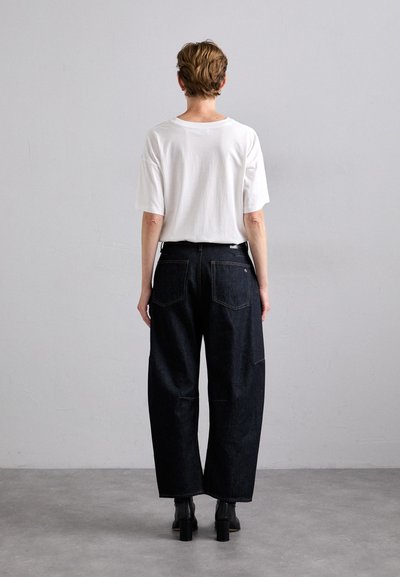 White short-sleeve t-shirt paired with high-waisted, wide-leg dark denim jeans. Black heeled boots. Plain gray background.