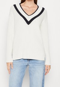 Kvinna i vit ribbad tröja med svarta V-formade ränder vid halslinningen och ljusblå denimjeans mot en neutral bakgrund.