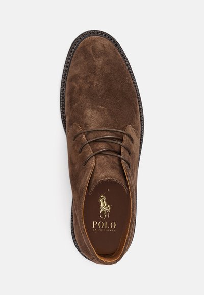 Zapato de ante marrón con diseño de cordones, punta redondeada, suela marrón y logo dorado de Polo Ralph Lauren en la plantilla. Detalle texturizado en la parte superior.