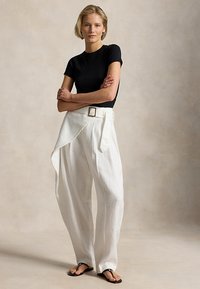 Γυναίκα που στέκεται με τα χέρια σταυρωμένα, φοράει μαύρο τοπ με κοντά μανίκια, λευκό παντελόνι με φαρδιά πόδια και ζώνη, και μαύρα σανδάλια σε ουδέτερο φόντο.