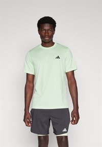 Hellgrünes Sport-T-Shirt mit kurzen Ärmeln und rundem Ausschnitt, ausgestattet mit einem kleinen schwarzen Adidas-Logo. Kombiniert mit dunklen Shorts.