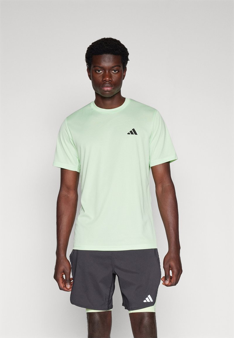 Hellgrünes Sport-T-Shirt mit kurzen Ärmeln und rundem Ausschnitt, ausgestattet mit einem kleinen schwarzen Adidas-Logo. Kombiniert mit dunklen Shorts.