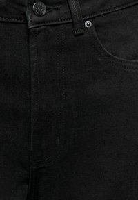 Detailní záběr na černé džínové kalhoty ukazující přední knoflík, poutko na opasek, nýtek a detaily stehu kapsy.