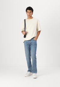 T-shirt à manches courtes crème, jeans droit bleu clair, baskets blanches, tenant un sac à bandoulière noir, se tenant devant un arrière-plan uni.