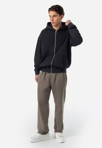 Moleton preto com fecho, de textura suave, com capuz, bolsos laterais e punhos canelados. Combinado com calças cinzas de corte solto e sapatilhas brancas.