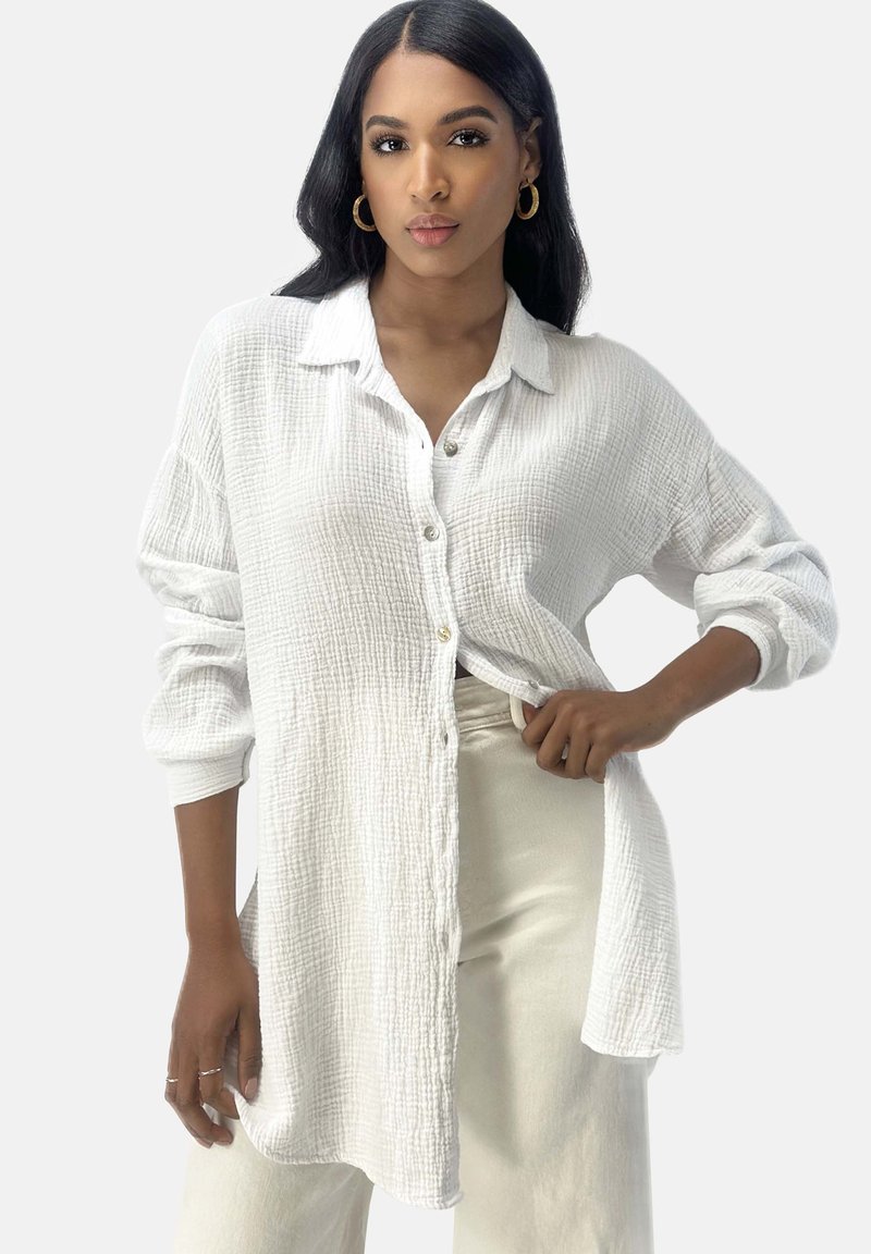 Chemise blanche à boutons en tissu texturé avec une finition froissée. Comprend des manches longues et un col, associée à un pantalon de couleur claire.