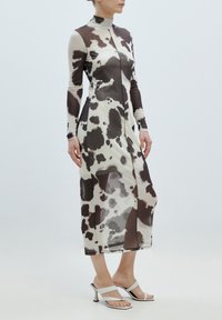 Langes, tailliertes Kleid mit einem transparenten Rinderhaut-Muster in Erdtönen. Es hat einen hohen Kragen und lange Ärmel, kombiniert mit weißen, hohen Sandalen.