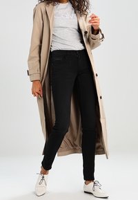 Trench beige sobre una camiseta gris con texto blanco, jeans ajustados negros y zapatos blancos de cordones. Tela suave, corte a medida.