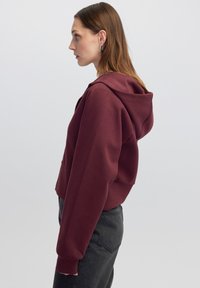 Sudadera corta color burdeos con capucha y bolsillo tipo canguro, con puños acanalados. Lleva puesta con vaqueros oscuros de cintura alta.