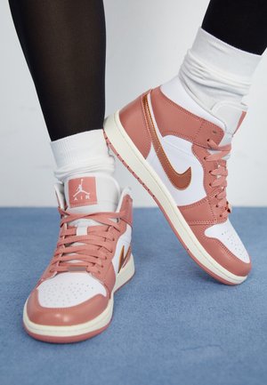 Personne portant des baskets Nike Air Jordan blanches et roses, un legging noir et des chaussettes blanches, debout sur un sol bleu.