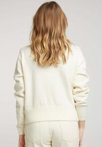 Pull en tricot couleur crème avec des poignets et un ourlet côtelés, présentant un design simple et une texture douce. Peut être associé à un pantalon de couleur claire.