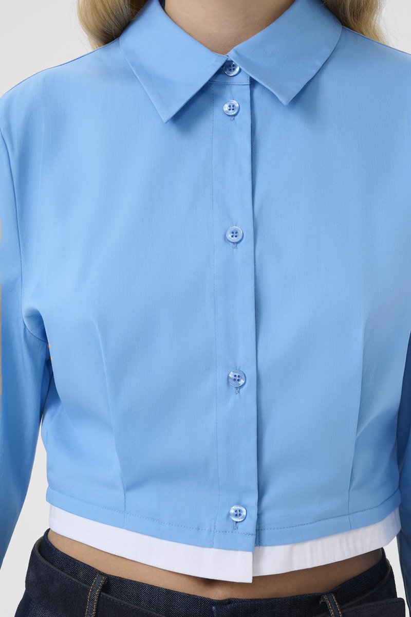 Femme portant une chemise courte bleu clair à boutons avec col, sur un débardeur blanc, associée à un jean bleu foncé.