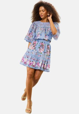 Frau in einem hellblauen Blumenkleid mit Puffärmeln und gerafftem Saum, die posiert und mit der Hand ihr Haar berührt, sie trägt beige Sandalen.