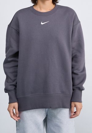 Personne portant un sweat-shirt ample gris foncé Nike avec un logo swoosh blanc centré sur la poitrine et un jean gris clair.