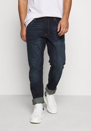 Mand iført mørkeblå bukser med op-rullede ben, hvide sneakers og en hvid skjorte, stående mod en ensartet lys baggrund.