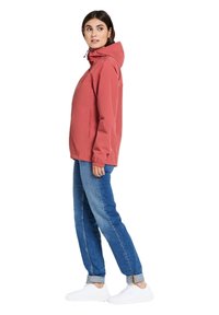 Rode capuchonjack van lichtgewicht stof. Voorzien van elastische manchetten en twee zijzakken. Gecombineerd met blauwe jeans en witte sneakers.