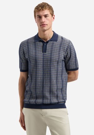 Junger Mann, der ein kurzärmeliges marineblau-beiges kariertes Poloshirt mit beigen Hosen trägt und vor einem weißen Hintergrund steht.