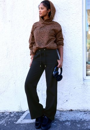 Brauner Kapuzenpullover mit Leopardenmuster, schwarze weite Hose und schwarze Sneakers. Das Modell hält Kopfhörer und steht vor einer strukturierten weißen Wand.