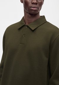 Homme portant un polo à manches longues vert olive avec deux boutons sur un fond clair uni.