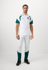 Άνδρας που στέκεται, φορώντας ιταλική ποδοσφαιρική στολή σε λευκό και πράσινο με λογότυπα, και μαύρα παπούτσια, σε απλό λευκό φόντο.