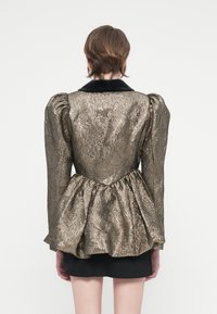 Metallik kuldne peplum jakk puhv-särkidega ja musta sametist kaelusega. Tekstureeritud pind lainelise mustriga. Kuvatud tagantvaade.