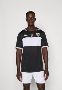Maillot de sport noir et blanc à manches courtes, avec une large bande blanche horizontale, des écussons de logo et un design texturé.