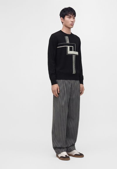 Jeune homme portant une chemise noire à manches longues avec un imprimé géométrique, un pantalon large gris à fines rayures et des chaussures décontractées bicolores sur un fond uni.