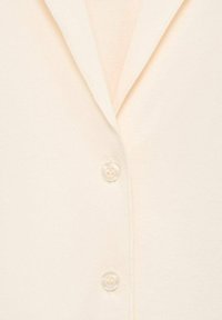 Button-up shirt dengan kain beige muda yang halus, menampilkan kerah runcing dan kancing plastik transparan.