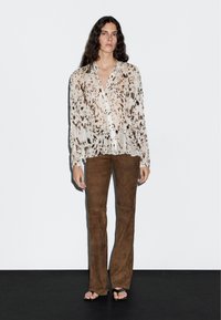 Blouse crème translucide avec un imprimé animalier marron, texture plissée et devant à boutons ; associée à un pantalon évasé en daim marron foncé et des sandales.