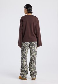 Braunes langärmeliges Oberteil kombiniert mit leichten, lockeren Hose im Tarnmuster in Grün, Grau und Beige. Braune Schuhe runden das Outfit ab.