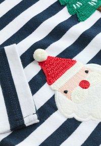Tengerészkék és fehér csíkos anyagon egy hímzett Mikulás-arc látható, piros sapkával, fehér szakállal és fekete szemekkel. Puha tapintású, pom-pom részletekkel.