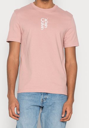 Mann trägt ein hellrosa Calvin Klein T-Shirt mit vertikalem weißen Logo-Schriftzug auf der Brust und hellblaue Jeans.